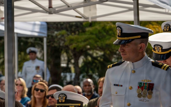 A formal U.S. Navy military ceremony is taking place outdoors under a white canopy tent. Several naval officers in white dress uniforms are present, along with a large seated audience of both military personnel and civilians. The event appears to be a significant ceremonial occasion such as a change of command, commissioning, or memorial service.