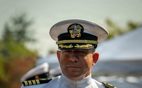 A U.S. Navy officer in full dress white uniform is rendering a salute at what appears to be a formal military ceremony. The officer displays an extensive array of medals, ribbons, and insignia on the right chest, indicating significant service and commendations. The white cap with gold oak leaf scrambled eggs on the brim suggests a senior officer rank of Captain (O-6) or above.