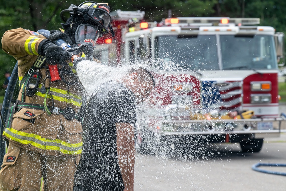 A fully equipped firefighter in turnout gear and SCBA mask is operating a fire hose, with water spraying forcefully toward the camera. A second person in civilian clothing appears to be getting soaked by the water stream, possibly during a training exercise or demonstration. A red and white fire engine with an American flag decal is visible in the background.