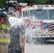 A fully equipped firefighter in turnout gear and SCBA mask is operating a fire hose, with water spraying forcefully toward the camera. A second person in civilian clothing appears to be getting soaked by the water stream, possibly during a training exercise or demonstration. A red and white fire engine with an American flag decal is visible in the background.