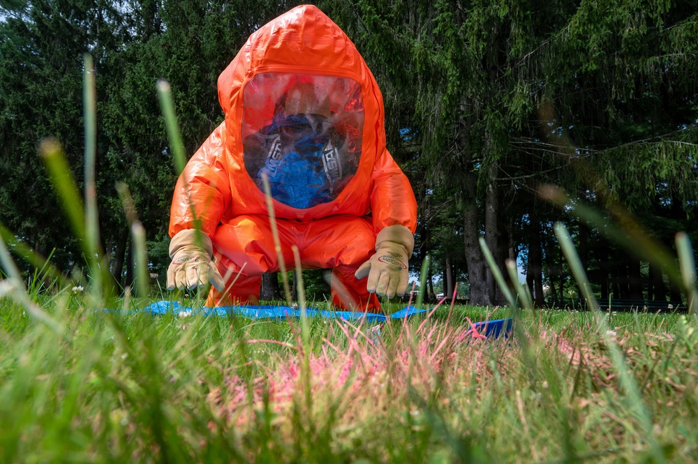 A person wearing a full orange hazmat (HAZMAT) suit with a blue respirator mask is kneeling on grass, appearing to examine or collect samples from the ground. The photo is taken from a ground-level perspective, emphasizing the grass in the foreground. Pink and blue markers or tape are visible on the grass near the subject, suggesting a controlled investigation or training exercise.