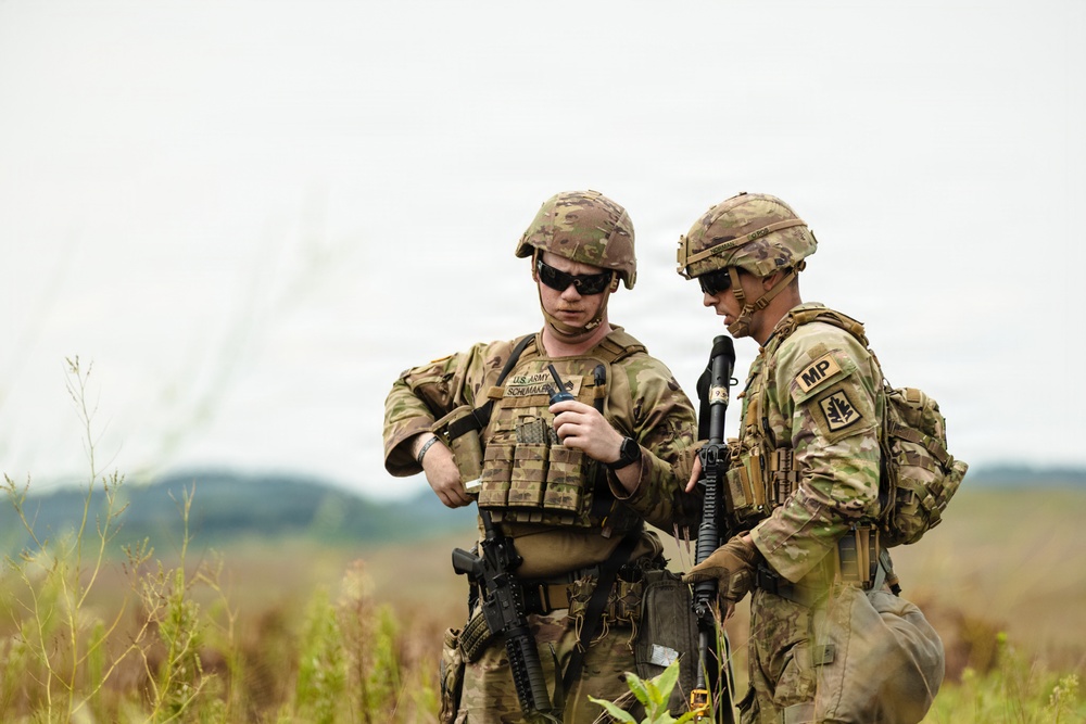Two U.S. Army soldiers in full combat gear are standing in an open field, appearing to consult a handheld device or document. Both wear MultiCam (OCP) camouflage uniforms, tactical vests, and helmets. The soldier on the right displays an 'MP' (Military Police) patch on his left shoulder.