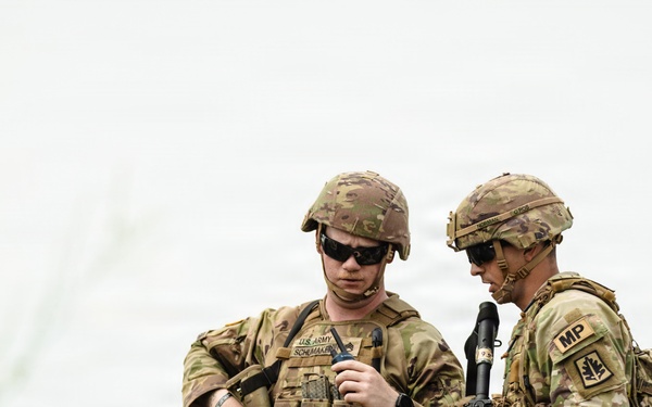 Two U.S. Army soldiers in full combat gear are standing in an open field, appearing to consult a handheld device or document. Both wear MultiCam (OCP) camouflage uniforms, tactical vests, and helmets. The soldier on the right displays an 'MP' (Military Police) patch on his left shoulder.