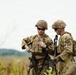 Two U.S. Army soldiers in full combat gear are standing in an open field, appearing to consult a handheld device or document. Both wear MultiCam (OCP) camouflage uniforms, tactical vests, and helmets. The soldier on the right displays an 'MP' (Military Police) patch on his left shoulder.
