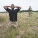 The image depicts what appears to be a military training exercise in an open field. A man in a black shirt and camouflage pants is kneeling with his hands behind his head in a surrender or detainee role-play position. In the background, a fully equipped soldier in camouflage uniform and helmet stands guard holding a rifle.