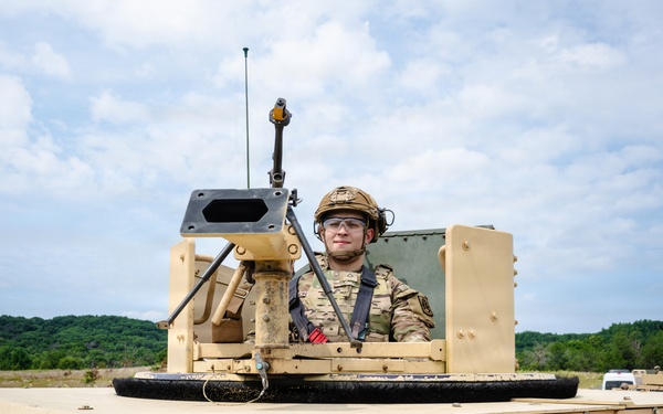 A soldier in full combat gear is positioned in the turret hatch of a tan/desert-colored HMMWV (Humvee) military vehicle. The soldier is wearing MultiCam/OCP camouflage uniform, a tactical helmet, and protective eyewear. The setting appears to be a training exercise in a rural, open field environment with tree lines visible in the background.