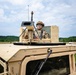 A soldier in full combat gear is positioned in the turret hatch of a tan/desert-colored HMMWV (Humvee) military vehicle. The soldier is wearing MultiCam/OCP camouflage uniform, a tactical helmet, and protective eyewear. The setting appears to be a training exercise in a rural, open field environment with tree lines visible in the background.