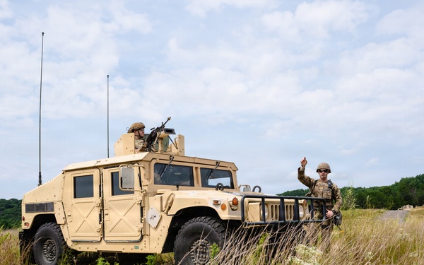 A military training exercise is underway in a rural, open field environment featuring at least two HMMWV (Humvee) vehicles on a dirt track. One soldier is positioned in the turret of the lead vehicle while another stands at the front, raising his hand in a signaling gesture. Additional vehicles and personnel are visible in the background.