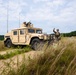 A military training exercise is underway in a rural, open field environment featuring at least two HMMWV (Humvee) vehicles on a dirt track. One soldier is positioned in the turret of the lead vehicle while another stands at the front, raising his hand in a signaling gesture. Additional vehicles and personnel are visible in the background.