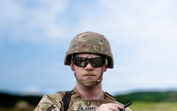 A U.S. Army soldier in full combat gear stands in an outdoor field environment. The soldier is wearing Operational Camouflage Pattern (OCP) uniform with a tactical vest, helmet, and ballistic sunglasses. A name tape reading 'SCHUMAN' and 'U.S. ARMY' is visible on the chest.