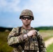A U.S. Army soldier in full combat gear stands in an outdoor field environment. The soldier is wearing Operational Camouflage Pattern (OCP) uniform with a tactical vest, helmet, and ballistic sunglasses. A name tape reading 'SCHUMAN' and 'U.S. ARMY' is visible on the chest.
