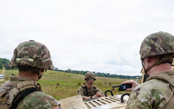 Three U.S. Army soldiers in OCP (Operational Camouflage Pattern) uniforms are gathered around a tan HMMWV (Humvee) in an open field training environment. One soldier is holding a paper shooting target with concentric circles, while another holds a radio device, and a third leans over the hood of the vehicle. A crew-served weapon is mounted on top of the Humvee.