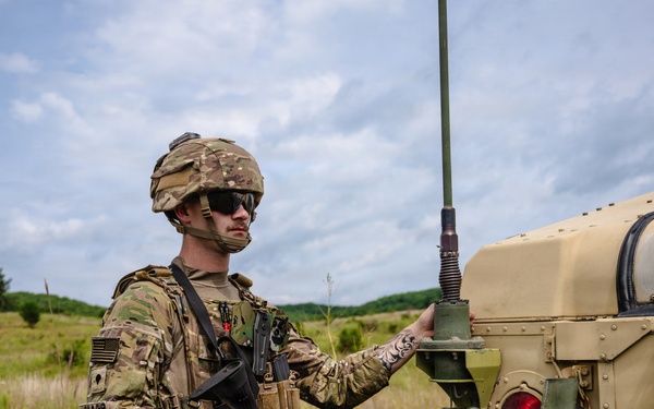 A soldier in full combat gear stands beside a tan military vehicle, likely a HMMWV (Humvee), in an open field training environment. The soldier is wearing MultiCam camouflage, a tactical vest, helmet with accessories, and is holding an M4-style carbine rifle. The setting appears to be a military training exercise based on the terrain and equipment configuration.
