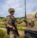 A soldier in full combat gear stands beside a tan military vehicle, likely a HMMWV (Humvee), in an open field training environment. The soldier is wearing MultiCam camouflage, a tactical vest, helmet with accessories, and is holding an M4-style carbine rifle. The setting appears to be a military training exercise based on the terrain and equipment configuration.
