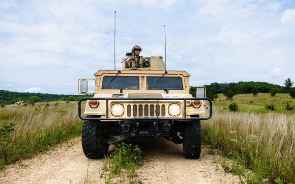 A military HMMWV (Humvee) is positioned on a dirt trail in an open grassy field, facing directly toward the camera. A soldier in full combat gear and helmet is visible standing through the roof hatch of the vehicle. The setting appears to be a training area or field exercise environment based on the terrain and vegetation.