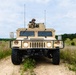 A military HMMWV (Humvee) is positioned on a dirt trail in an open grassy field, facing directly toward the camera. A soldier in full combat gear and helmet is visible standing through the roof hatch of the vehicle. The setting appears to be a training area or field exercise environment based on the terrain and vegetation.