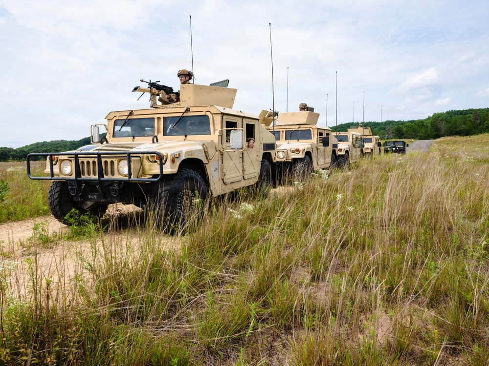 A convoy of military HMMWV (High Mobility Multipurpose Wheeled Vehicle) Humvees in desert tan paint scheme is traveling along a dirt trail through an open grassy field. At least four to five vehicles are visible in a column formation, with the lead vehicle equipped with a mounted crew-served weapon and a gunner in the turret. The setting appears to be a military training area based on the terrain and vegetation.