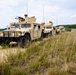 A convoy of military HMMWV (High Mobility Multipurpose Wheeled Vehicle) Humvees in desert tan paint scheme is traveling along a dirt trail through an open grassy field. At least four to five vehicles are visible in a column formation, with the lead vehicle equipped with a mounted crew-served weapon and a gunner in the turret. The setting appears to be a military training area based on the terrain and vegetation.