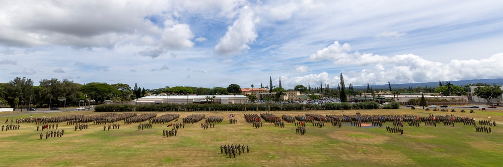A large military formation ceremony is taking place on an expansive parade ground, with hundreds of uniformed personnel arranged in precise rectangular unit formations across the field. The aerial/elevated perspective reveals multiple distinct unit blocks, color guards with flags, and what appears to be a formal review or parade event. The setting appears to be a military installation in a tropical environment, likely Hawaii based on the vegetation and terrain.