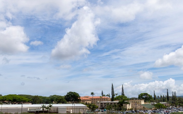 A large military formation ceremony is taking place on an expansive parade ground, with hundreds of uniformed personnel arranged in precise rectangular unit formations across the field. The aerial/elevated perspective reveals multiple distinct unit blocks, color guards with flags, and what appears to be a formal review or parade event. The setting appears to be a military installation in a tropical environment, likely Hawaii based on the vegetation and terrain.