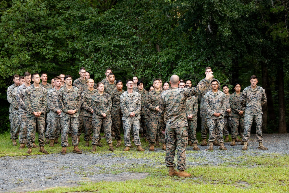 A military instructor or senior non-commissioned officer addresses a large formation of uniformed personnel in an outdoor wooded setting. The group, wearing matching camouflage uniforms consistent with U.S. Marine Corps MARPAT pattern, stands attentively while the speaker gestures with a raised hand. The setting appears to be a training area or field environment with gravel ground and dense tree cover in the background.