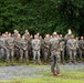 A military instructor or senior non-commissioned officer addresses a large formation of uniformed personnel in an outdoor wooded setting. The group, wearing matching camouflage uniforms consistent with U.S. Marine Corps MARPAT pattern, stands attentively while the speaker gestures with a raised hand. The setting appears to be a training area or field environment with gravel ground and dense tree cover in the background.