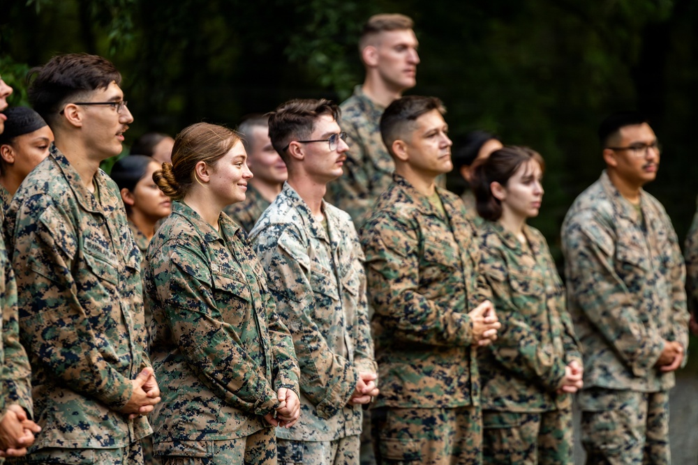 A group of military personnel in camouflage uniforms are standing in formation outdoors, appearing to be at attention or listening to a presentation or ceremony. The individuals are wearing what appears to be U.S. Marine Corps MARPAT (Marine Pattern) digital camouflage uniforms. The group is diverse in gender and ethnicity, standing in rows against a backdrop of green foliage.