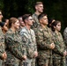A group of military personnel in camouflage uniforms are standing in formation outdoors, appearing to be at attention or listening to a presentation or ceremony. The individuals are wearing what appears to be U.S. Marine Corps MARPAT (Marine Pattern) digital camouflage uniforms. The group is diverse in gender and ethnicity, standing in rows against a backdrop of green foliage.