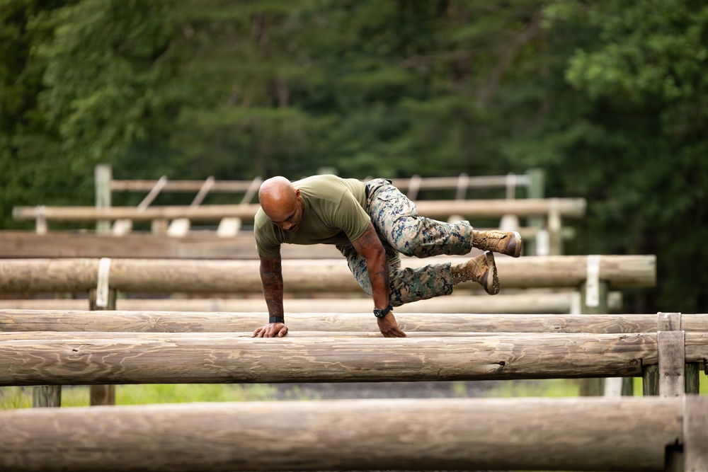 A military service member in USMC digital camouflage utilities and combat boots is navigating a log obstacle course, appearing to vault or crawl over a series of horizontal logs. The individual is bald, wearing a green t-shirt, and has visible tattoos on their arms. The setting is an outdoor military training facility surrounded by dense green trees.