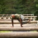 A military service member in USMC digital camouflage utilities and combat boots is navigating a log obstacle course, appearing to vault or crawl over a series of horizontal logs. The individual is bald, wearing a green t-shirt, and has visible tattoos on their arms. The setting is an outdoor military training facility surrounded by dense green trees.