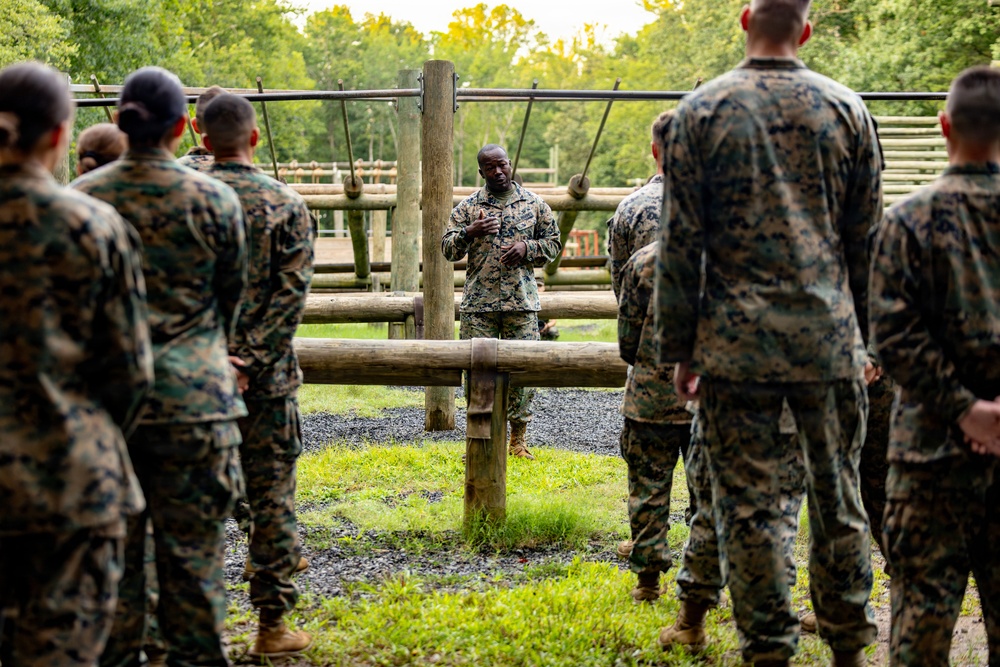 A military instructor or senior non-commissioned officer addresses a group of personnel in woodland camouflage uniforms at an outdoor obstacle course. The speaker stands facing the group, gesturing with his hands, while the others listen attentively with their backs to the camera. The setting appears to be a military training facility surrounded by trees.