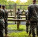 A military instructor or senior non-commissioned officer addresses a group of personnel in woodland camouflage uniforms at an outdoor obstacle course. The speaker stands facing the group, gesturing with his hands, while the others listen attentively with their backs to the camera. The setting appears to be a military training facility surrounded by trees.