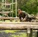 Military personnel in camouflage uniforms are navigating a wooden log obstacle course in an outdoor training environment. The foreground shows one soldier actively vaulting over parallel horizontal logs, while several others are visible in the background performing similar exercises. A larger rope and timber obstacle structure is visible in the background, surrounded by dense green forest.