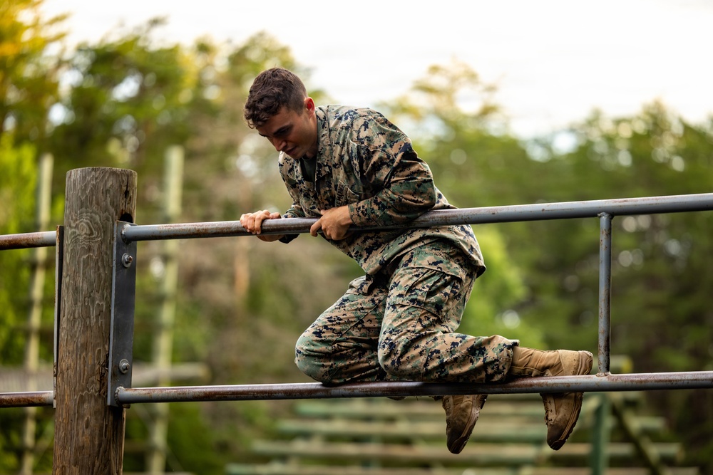 A young male service member in MARPAT (Marine digital camouflage) uniform is navigating an obstacle course, specifically climbing over a horizontal metal bar structure. He is mid-movement, gripping the top rail while pulling his body over it. The background shows a wooded outdoor training environment with additional obstacle course elements visible.