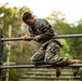 A young male service member in MARPAT (Marine digital camouflage) uniform is navigating an obstacle course, specifically climbing over a horizontal metal bar structure. He is mid-movement, gripping the top rail while pulling his body over it. The background shows a wooded outdoor training environment with additional obstacle course elements visible.