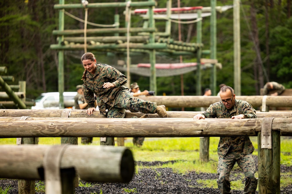 Two military personnel in camouflage uniforms are navigating a log obstacle course at what appears to be a military training facility. A female service member is climbing over horizontal logs while a male service member with glasses assists or navigates alongside her. Additional personnel and a larger obstacle course structure are visible in the background.