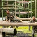 Two military personnel in camouflage uniforms are navigating a log obstacle course at what appears to be a military training facility. A female service member is climbing over horizontal logs while a male service member with glasses assists or navigates alongside her. Additional personnel and a larger obstacle course structure are visible in the background.