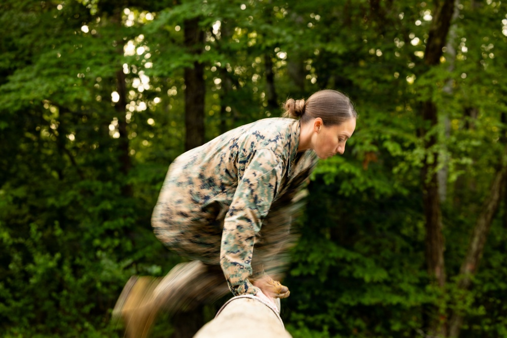 A person wearing military camouflage (MARPAT/digital pattern consistent with US Marine Corps uniform) is captured mid-motion vaulting or climbing over a large log or beam obstacle. The motion blur indicates rapid movement, suggesting this is part of a military obstacle course or physical training exercise. The setting is a wooded outdoor environment with dense green foliage in the background.