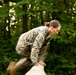 A person wearing military camouflage (MARPAT/digital pattern consistent with US Marine Corps uniform) is captured mid-motion vaulting or climbing over a large log or beam obstacle. The motion blur indicates rapid movement, suggesting this is part of a military obstacle course or physical training exercise. The setting is a wooded outdoor environment with dense green foliage in the background.