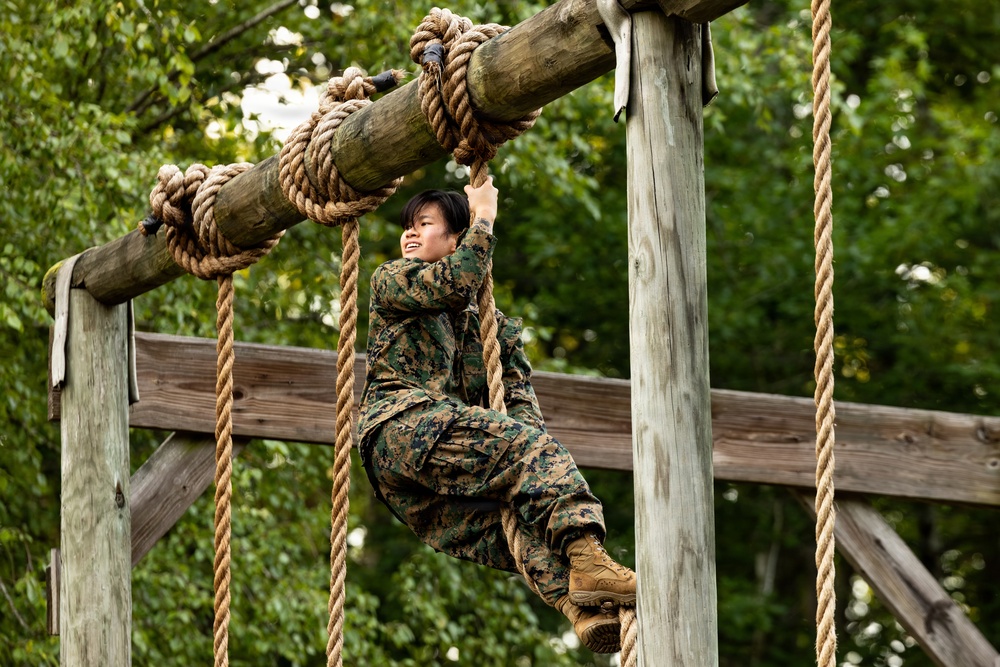A person in military camouflage uniform is navigating a rope obstacle course, gripping a hanging rope while suspended off the ground. The individual appears to be smiling or grimacing with effort, suggesting active physical exertion during what appears to be military training. The setting is an outdoor wooden obstacle course structure surrounded by green foliage.