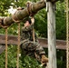 A person in military camouflage uniform is navigating a rope obstacle course, gripping a hanging rope while suspended off the ground. The individual appears to be smiling or grimacing with effort, suggesting active physical exertion during what appears to be military training. The setting is an outdoor wooden obstacle course structure surrounded by green foliage.