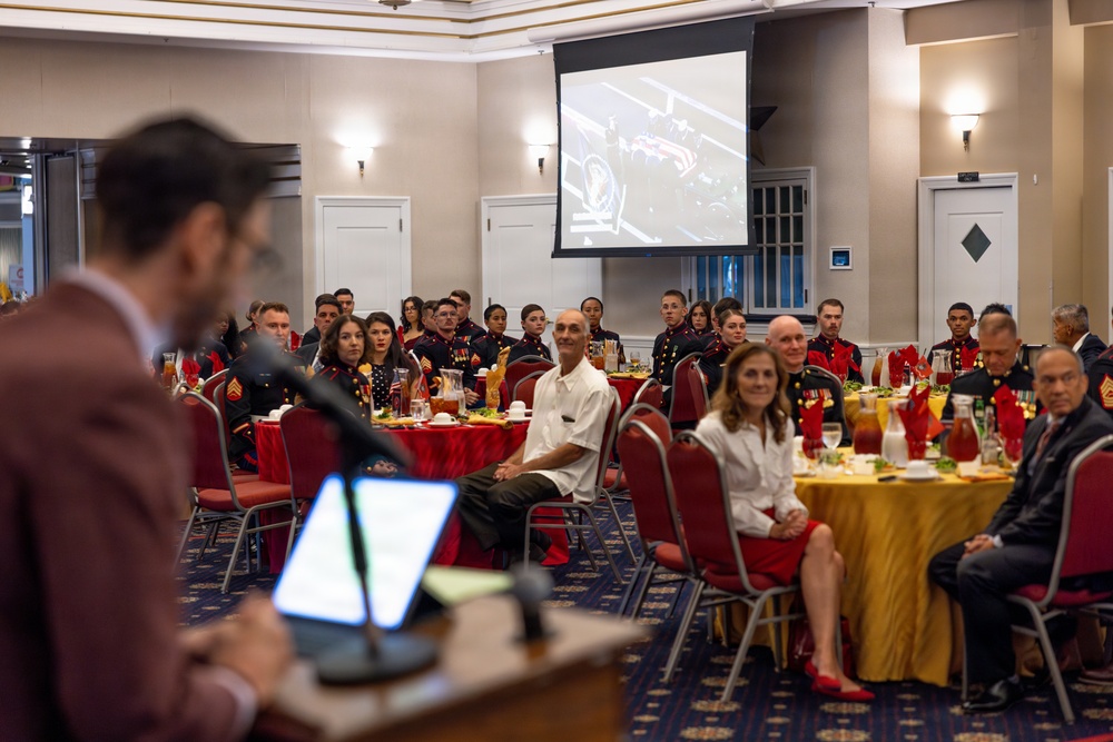A formal military banquet or awards ceremony is taking place in a decorated ballroom, with numerous U.S. Marines in dress blues seated at round tables with red and gold tablecloths. A speaker at a podium addresses the audience while a projection screen displays an image in the background. Civilians and military personnel are seated together, suggesting a mixed-audience formal event.
