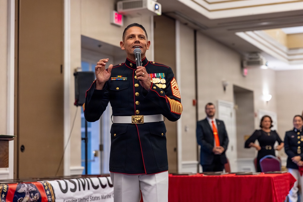A United States Marine Corps senior non-commissioned officer in dress blue uniform is speaking at a formal event, holding a microphone. The setting appears to be a banquet hall or ballroom, with a banner partially visible reading 'USMCCC' and 'Serving United States Marine Corps.' Several attendees in formal attire are visible in the background.