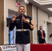 A United States Marine Corps senior non-commissioned officer in dress blue uniform is speaking at a formal event, holding a microphone. The setting appears to be a banquet hall or ballroom, with a banner partially visible reading 'USMCCC' and 'Serving United States Marine Corps.' Several attendees in formal attire are visible in the background.