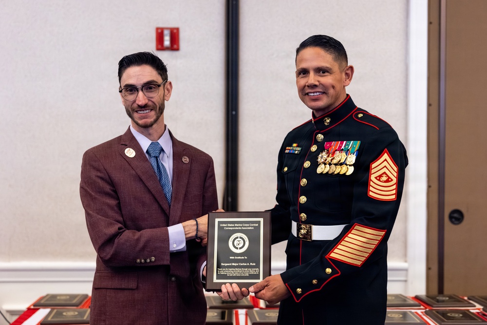 Two men pose together holding a plaque from the United States Marine Corps Combat Correspondents Association presented to Sergeant Major Carlos A. Ruiz. The man on the right is wearing a formal U.S. Marine Corps dress blue uniform with multiple medals, ribbons, and sergeant major chevrons. The event appears to be a formal military awards ceremony held in a banquet or conference room setting.