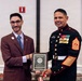 Two men pose together holding a plaque from the United States Marine Corps Combat Correspondents Association presented to Sergeant Major Carlos A. Ruiz. The man on the right is wearing a formal U.S. Marine Corps dress blue uniform with multiple medals, ribbons, and sergeant major chevrons. The event appears to be a formal military awards ceremony held in a banquet or conference room setting.