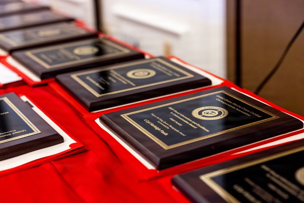 The image shows multiple military award plaques arranged on a red cloth-covered table. The plaques appear to be from the United States Marine Corps Combat Correspondents Association, featuring gold lettering and the Marine Corps seal on dark wood and black backgrounds. At least one plaque is legible enough to indicate a 'Visual Information and Excellence' award with 'First Place' designation.