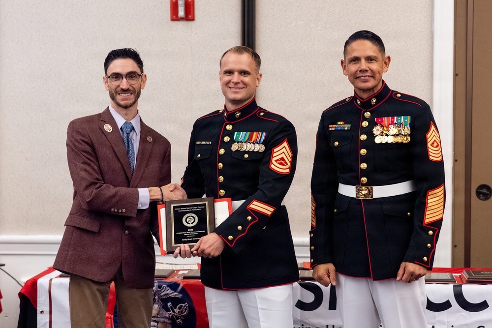 Three men pose for a formal photo at what appears to be a military awards ceremony. Two individuals are wearing United States Marine Corps dress blue uniforms with medals and rank insignia, while a civilian in a brown blazer shakes hands with one of the Marines while holding a plaque. The setting appears to be an indoor event venue with patriotic decorations visible in the background.
