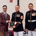 Three men pose for a formal photo at what appears to be a military awards ceremony. Two individuals are wearing United States Marine Corps dress blue uniforms with medals and rank insignia, while a civilian in a brown blazer shakes hands with one of the Marines while holding a plaque. The setting appears to be an indoor event venue with patriotic decorations visible in the background.
