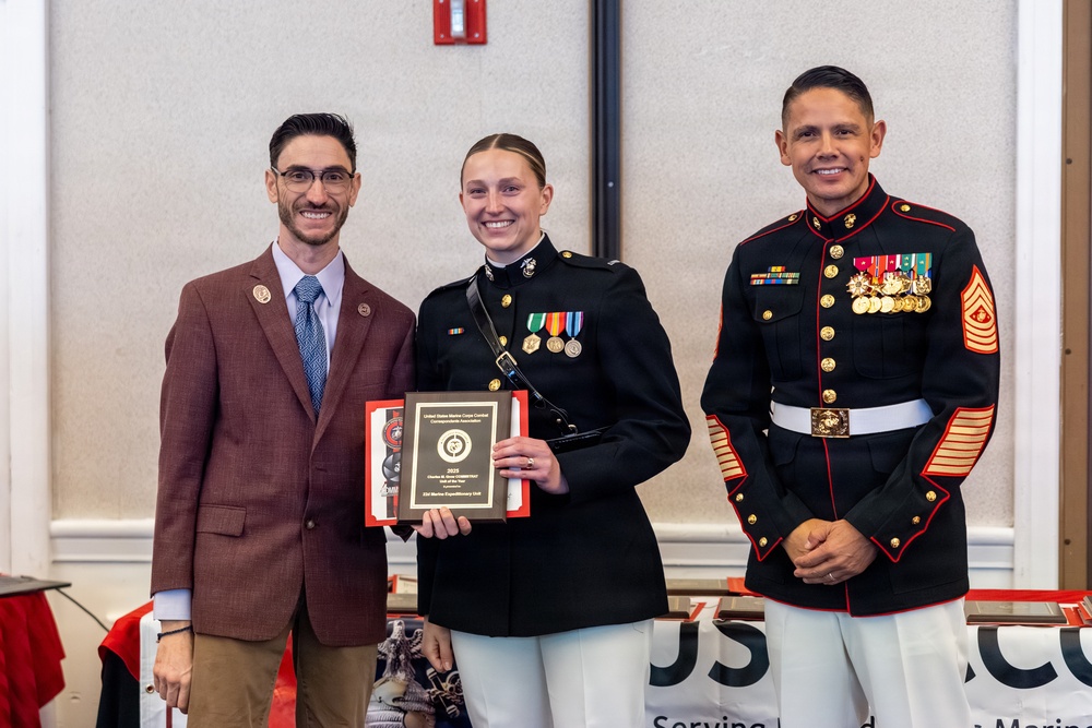 Three individuals pose for a formal photograph at what appears to be a military awards ceremony. A female Marine in dress blues holds a plaque award, flanked by a civilian in a brown suit and a senior enlisted Marine in dress blues with extensive medals and chevrons. The plaque appears to be from the United States Marine Corps Correspondents Association for the year 2023.